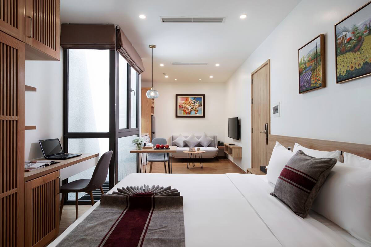 A modern hotel room with a large bed, a desk with a laptop, a sofa, and a TV.