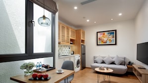 A modern living room with a sofa, a dining table with apples, a washing machine, and a kitchen area with wooden cabinets.