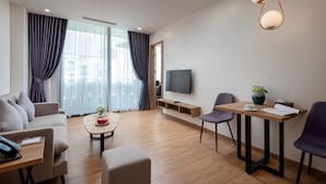 A modern hotel room with a sofa, a coffee table, a TV, and a dining area with chairs and a table.