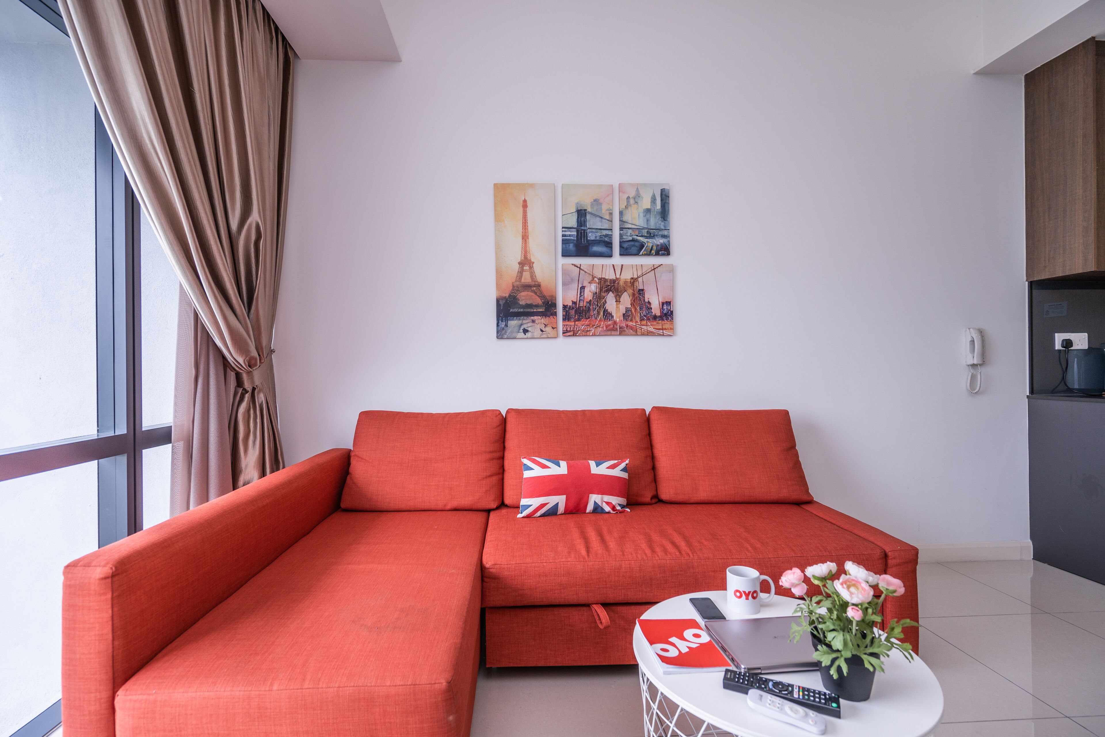 A modern living room with a red sofa, a small round table with a remote control, a smartphone, and a cup. There are two paintings on the wall depicting famous landmarks.