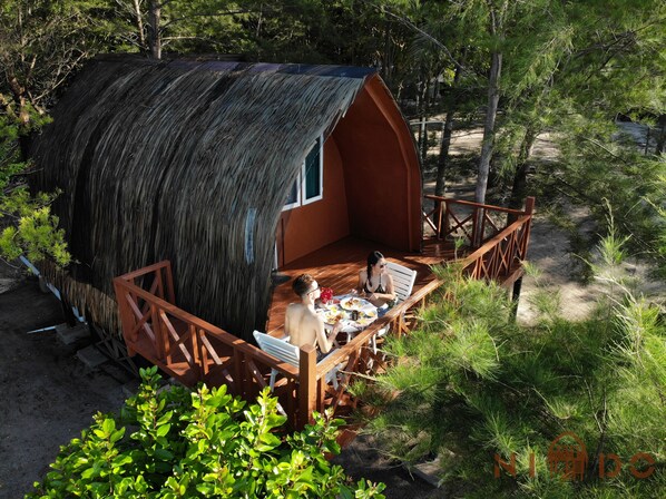 Une petite cabane à toit de chaume, dotée d’une terrasse en bois et d’une petite fenêtre, entourée de verdure.