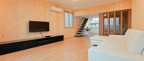 A modern living room with a flat-screen TV, a white sofa, wooden flooring, and a staircase leading to a kitchen area.