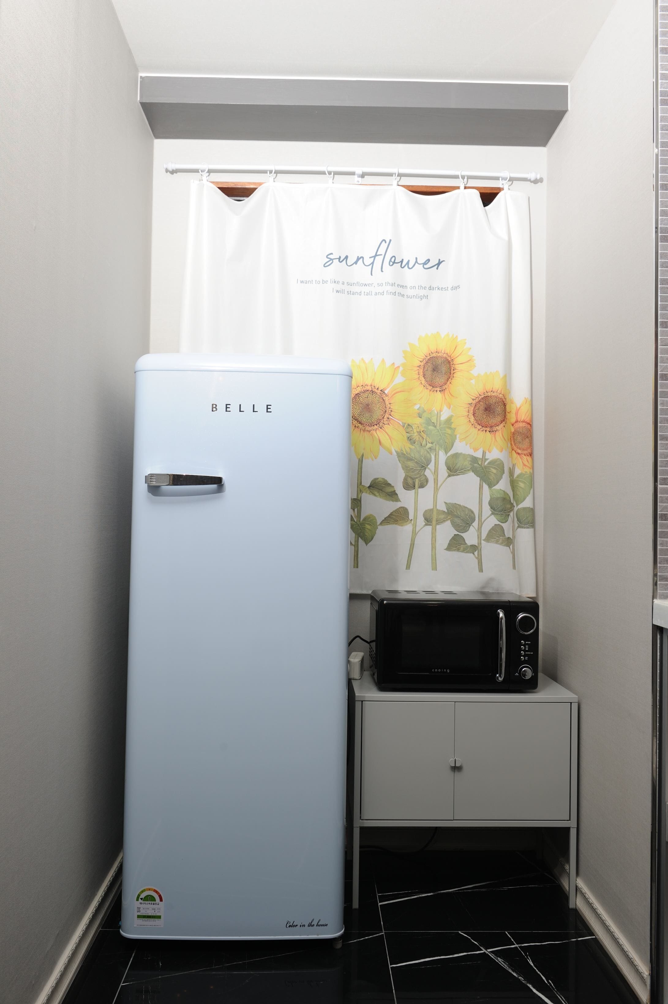 Een compact keukenhoekje met een lichtblauwe koelkast, een magnetron op een klein wit kastje, en een douchegordijn met zonnebloemen.