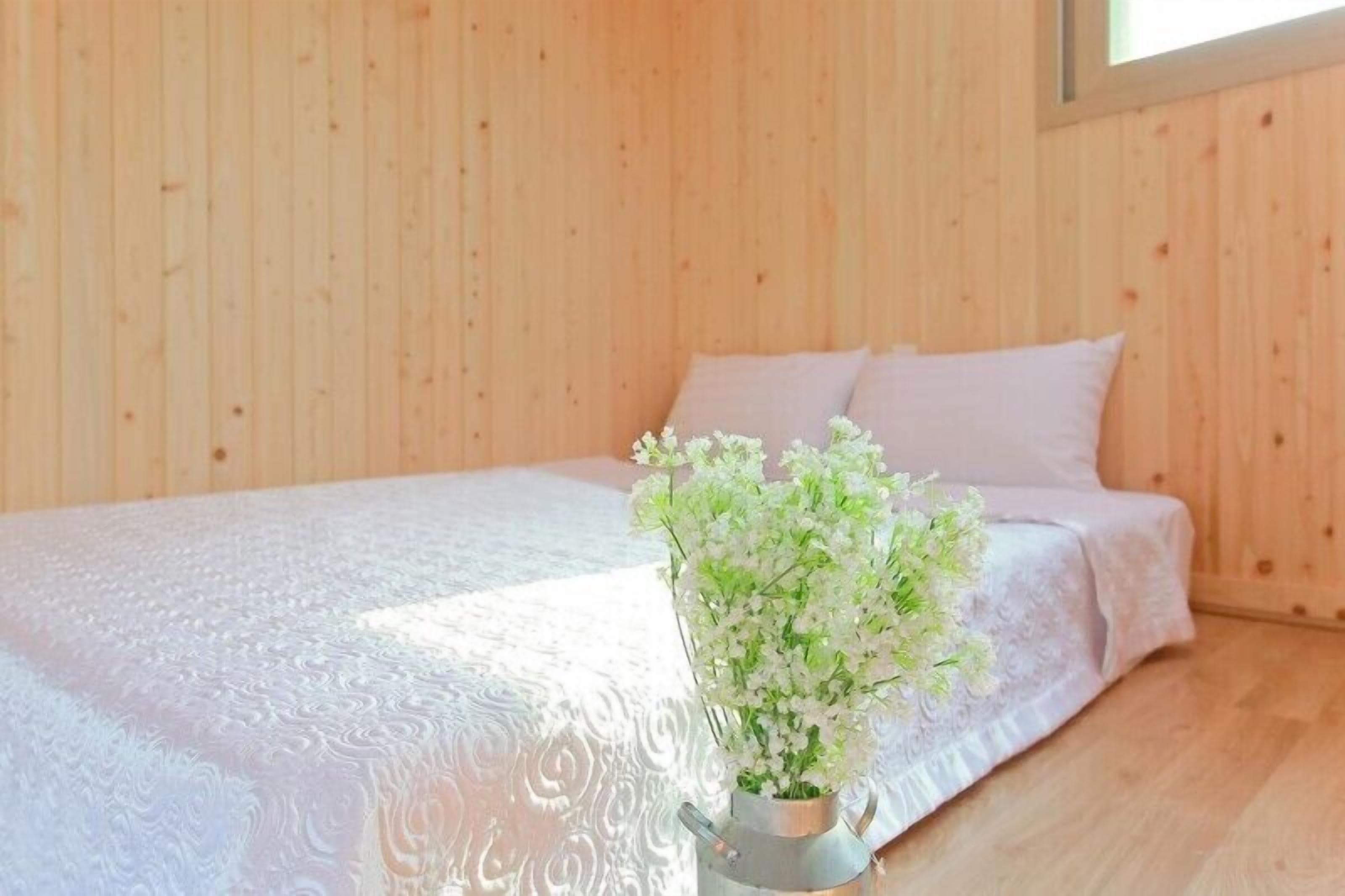 A bedroom with a wooden wall, a bed with white bedding, and a metal bucket with white flowers.