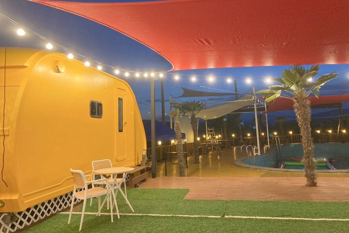 A yellow trailer-style room with a small table and chairs, a pool area with palm trees, and string lights.