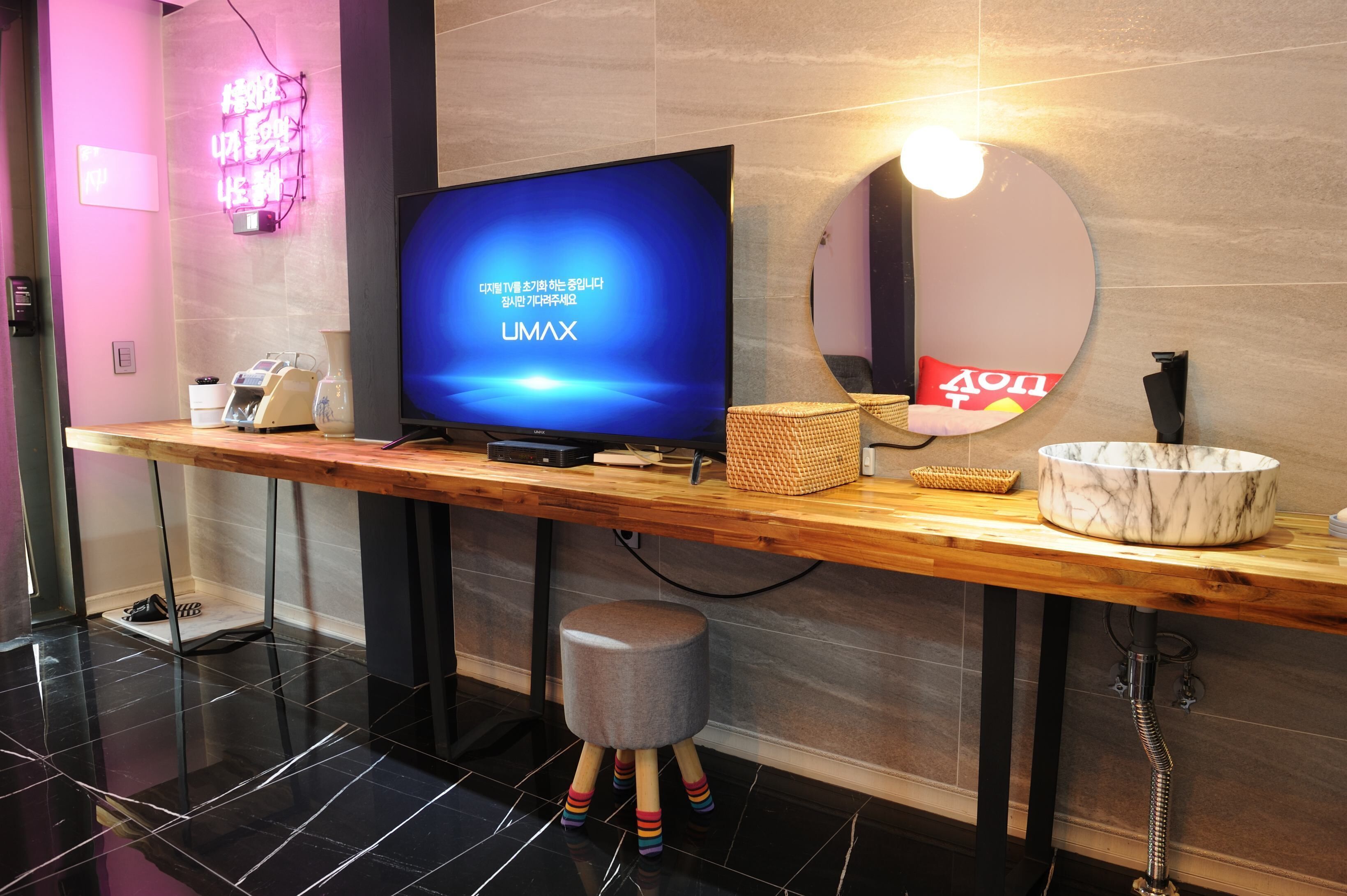 Ein moderner Hotel-Empfangsbereich mit einem Flachbildfernseher, der 'UMAX' anzeigt, einem hölzernen Empfangstresen, einer Marmorschale und einem runden Spiegel.