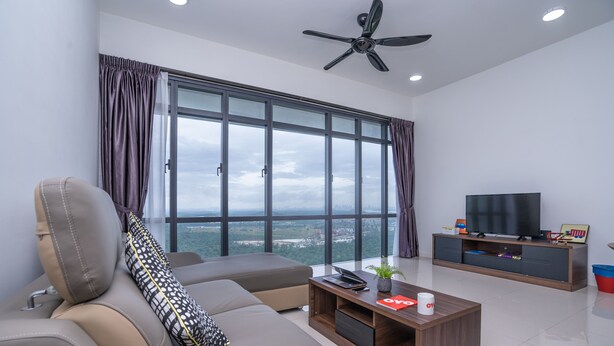 A modern living room with a sofa, coffee table, and TV, large windows with curtains, and a ceiling fan. - OYO Home 89725 Beautiful 3br Grand Medini (Iskandar Puteri)