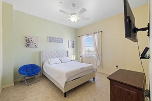 A bedroom with a bed, a ceiling fan, a window with curtains, and a small wooden cabinet.