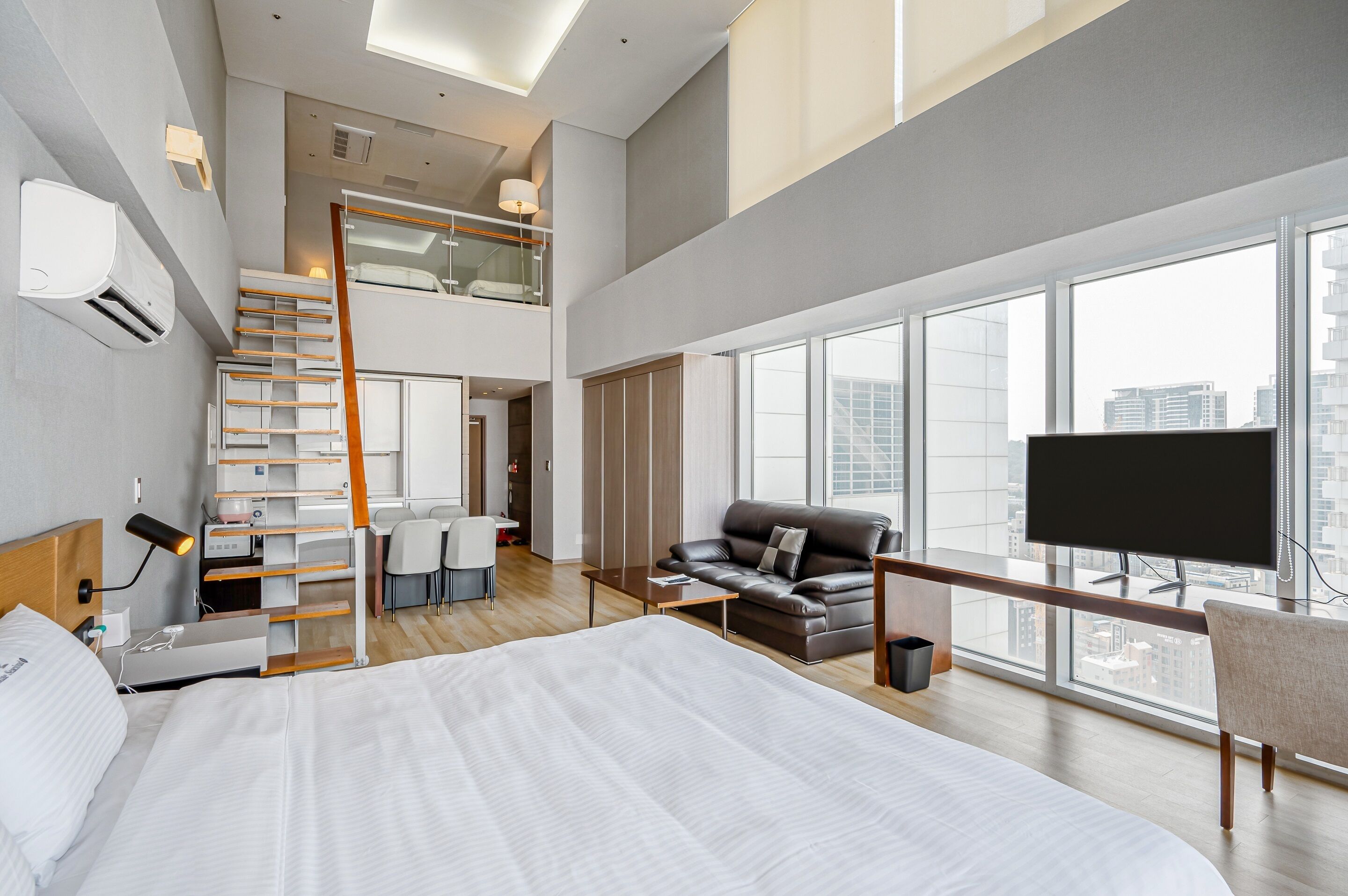 A modern hotel room with a large bed, a desk, a sofa, and a TV. There is a balcony with a city view.