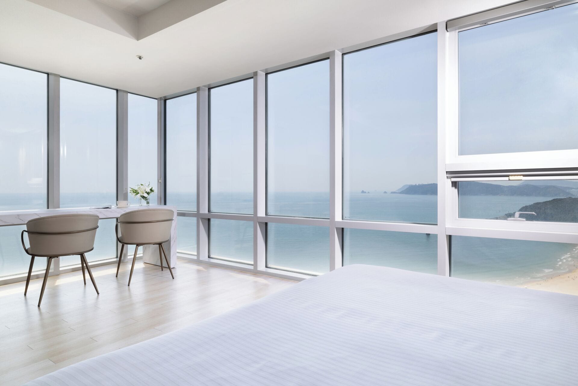 A modern bedroom with a large bed, two chairs, and a table, overlooking a beach and ocean.