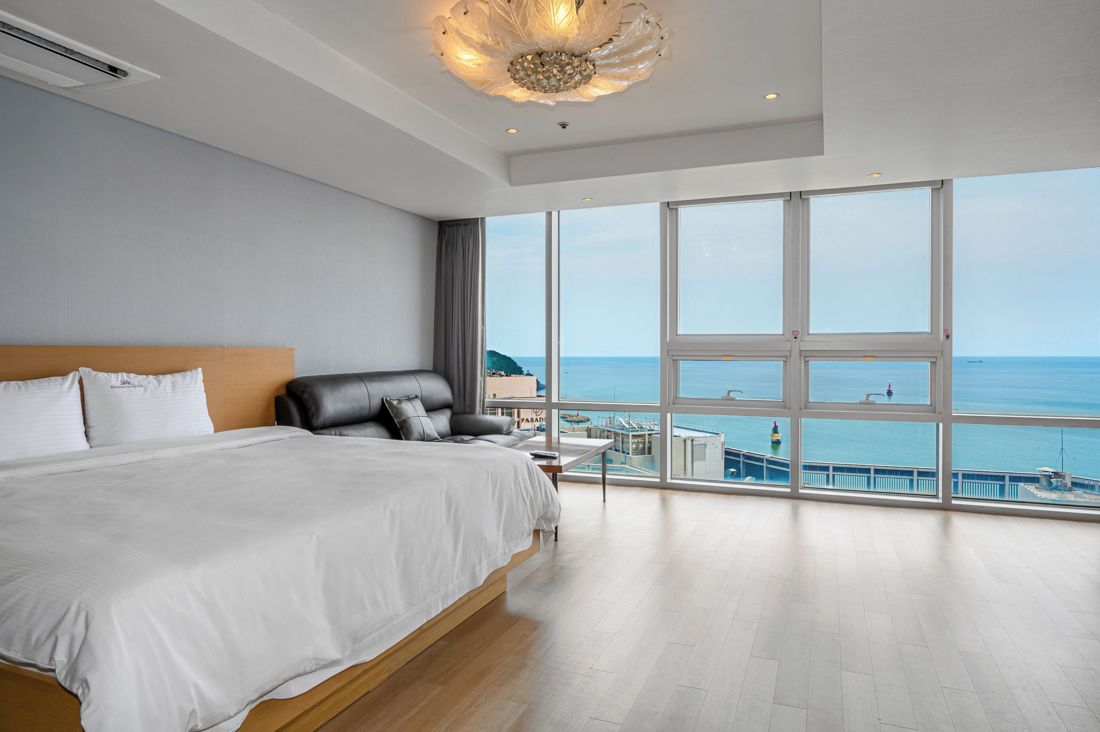Un grand lit avec une tête de lit en bois, une vue sur la mer grâce à de grandes fenêtres du sol au plafond et un lustre moderne.