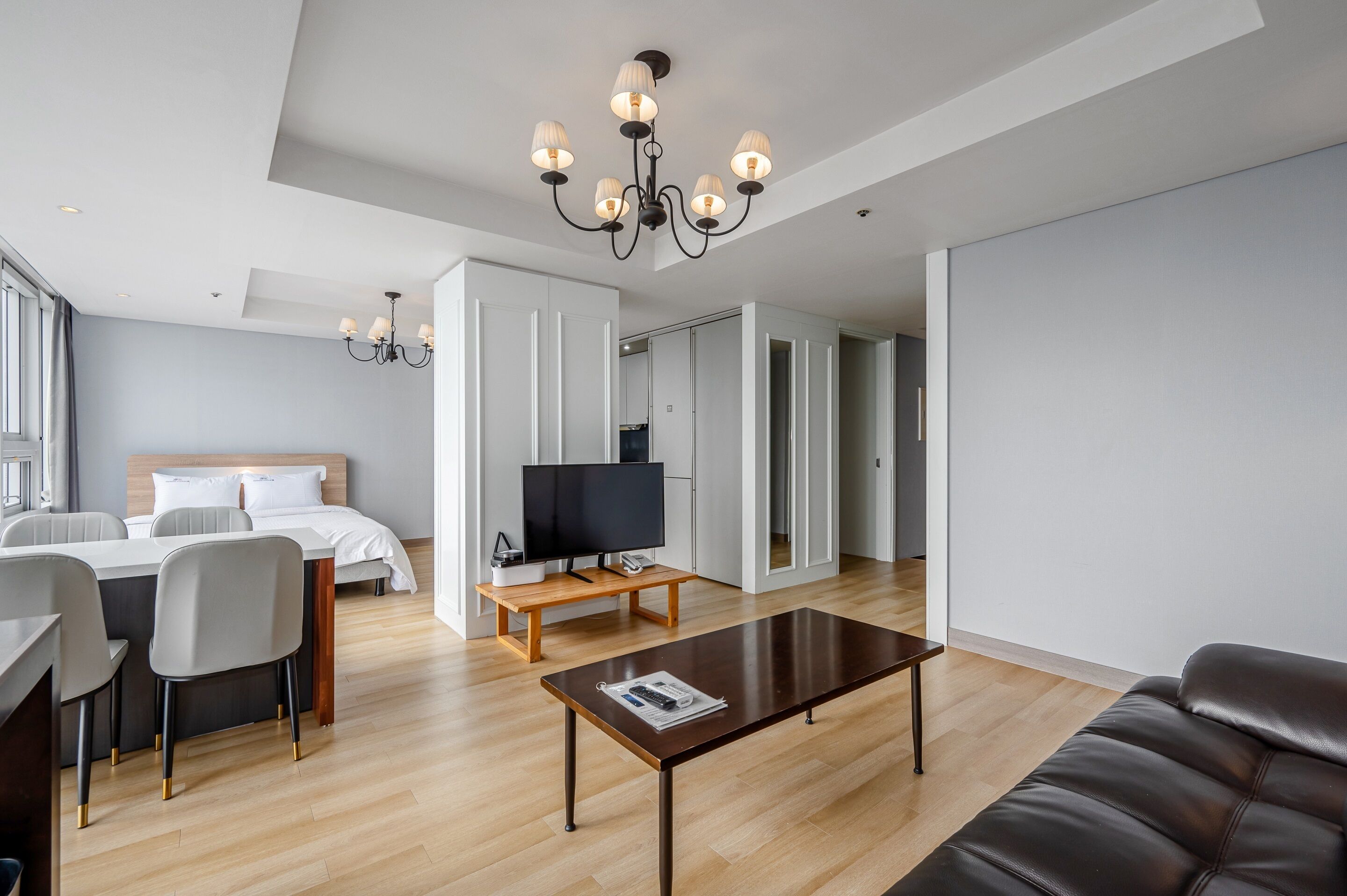 A modern hotel room with a bed, a TV on a wooden stand, a dining area with chairs, a coffee table, and a large mirror.