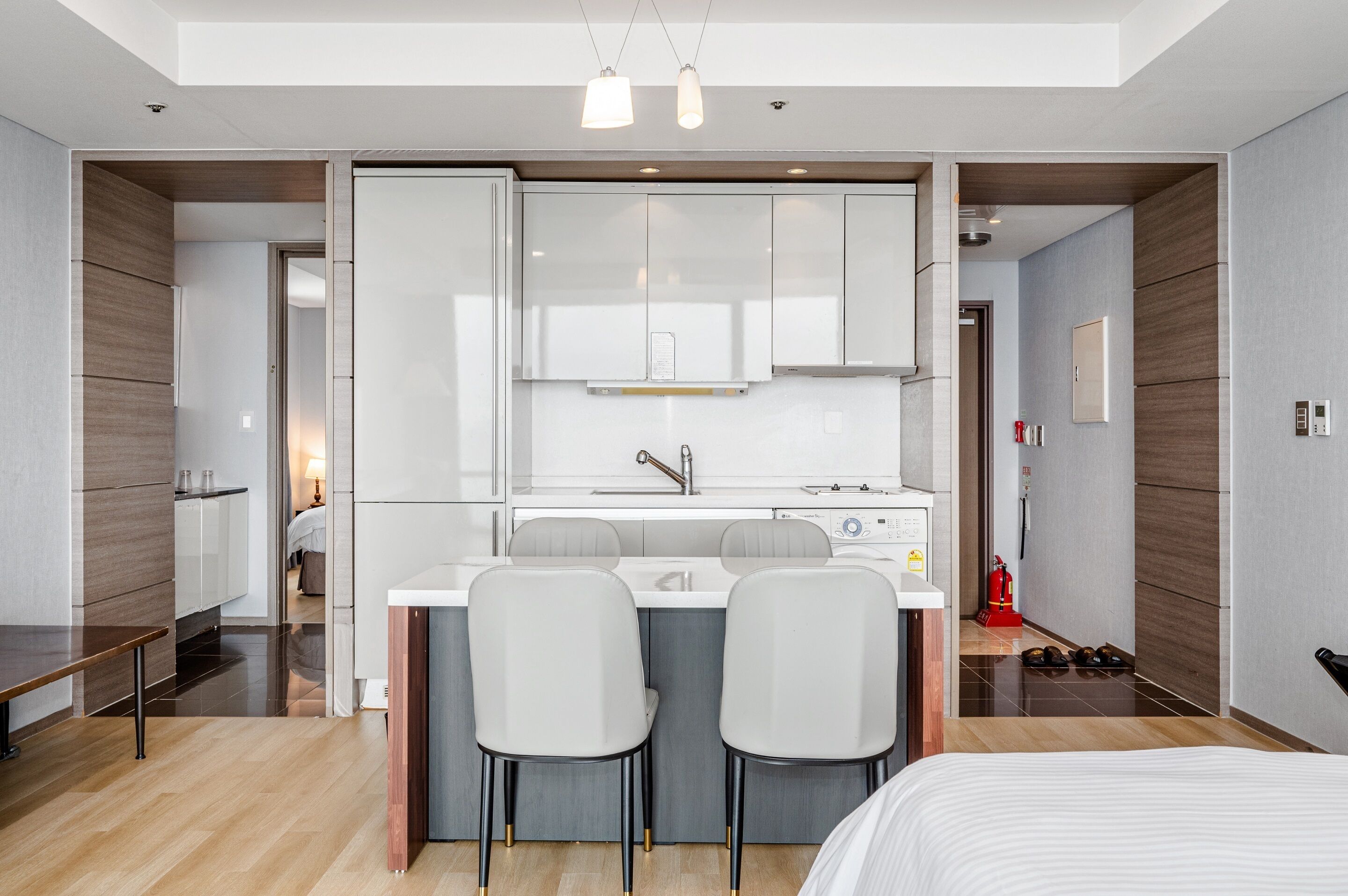 A modern hotel room with a kitchenette, two white bar stools, a bed, and a wooden floor.