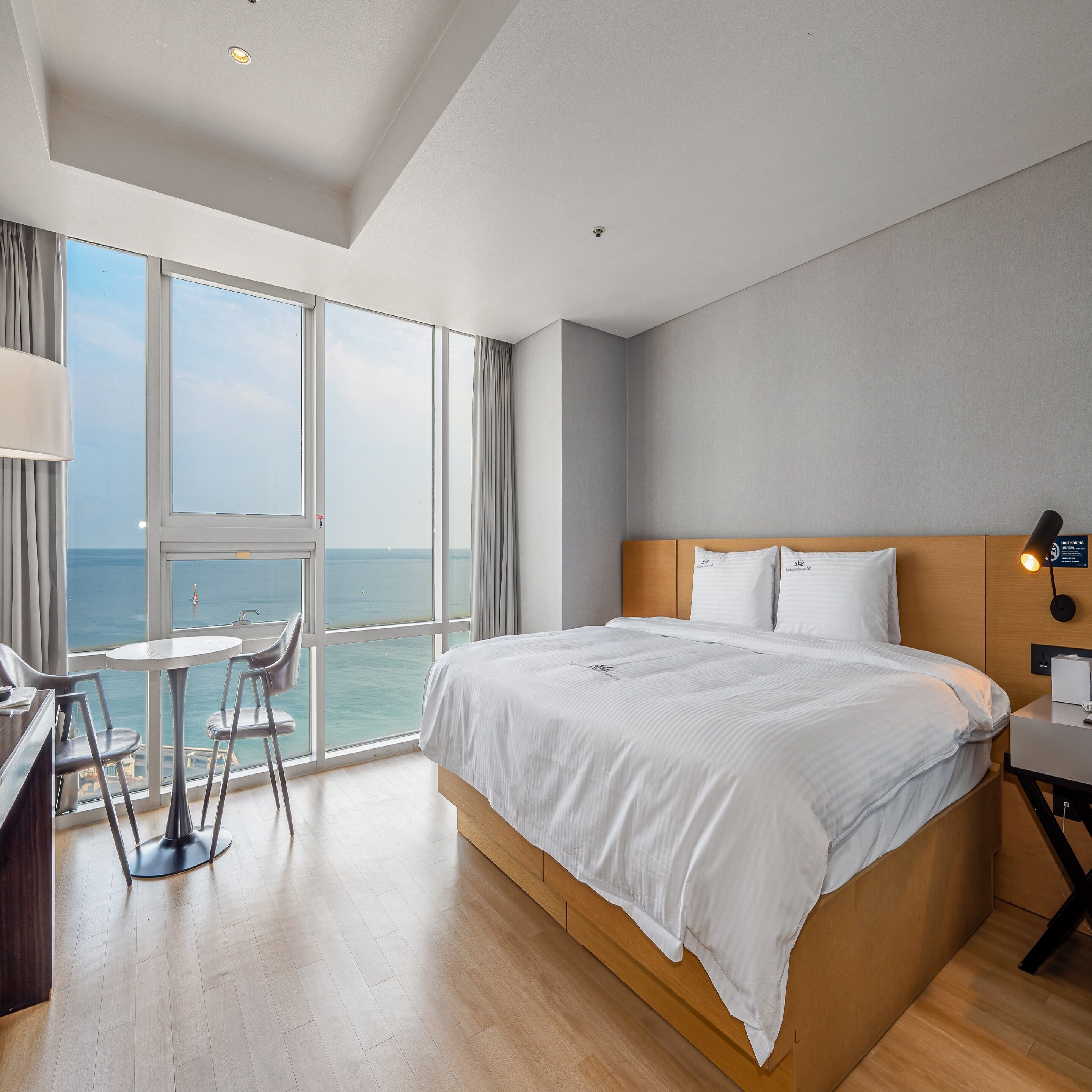 A hotel room with a large bed, a desk with a chair, and a view of the sea through large windows.