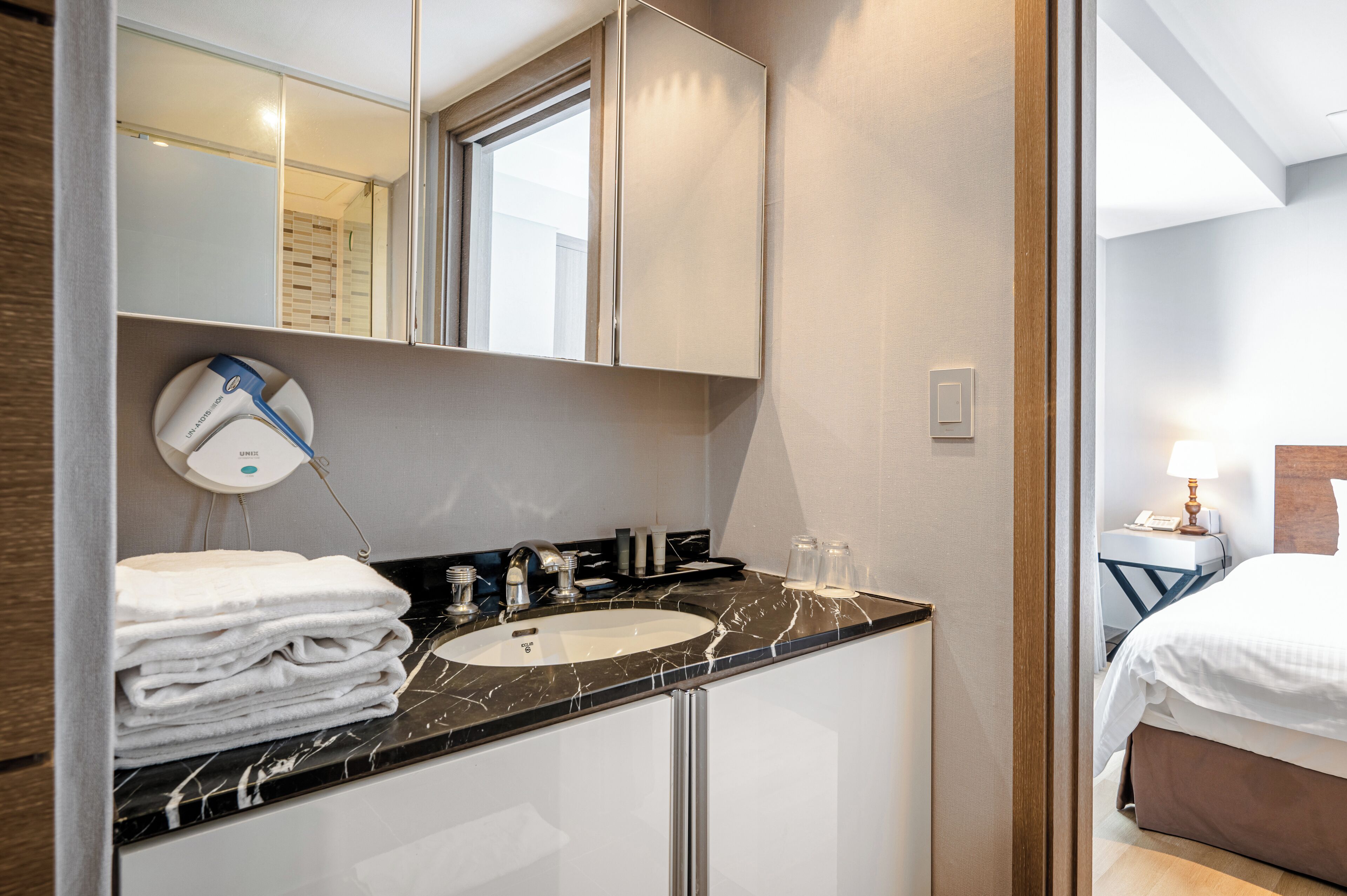A hotel bathroom with a sink, mirror, towel stack, and a nightstand with a lamp.