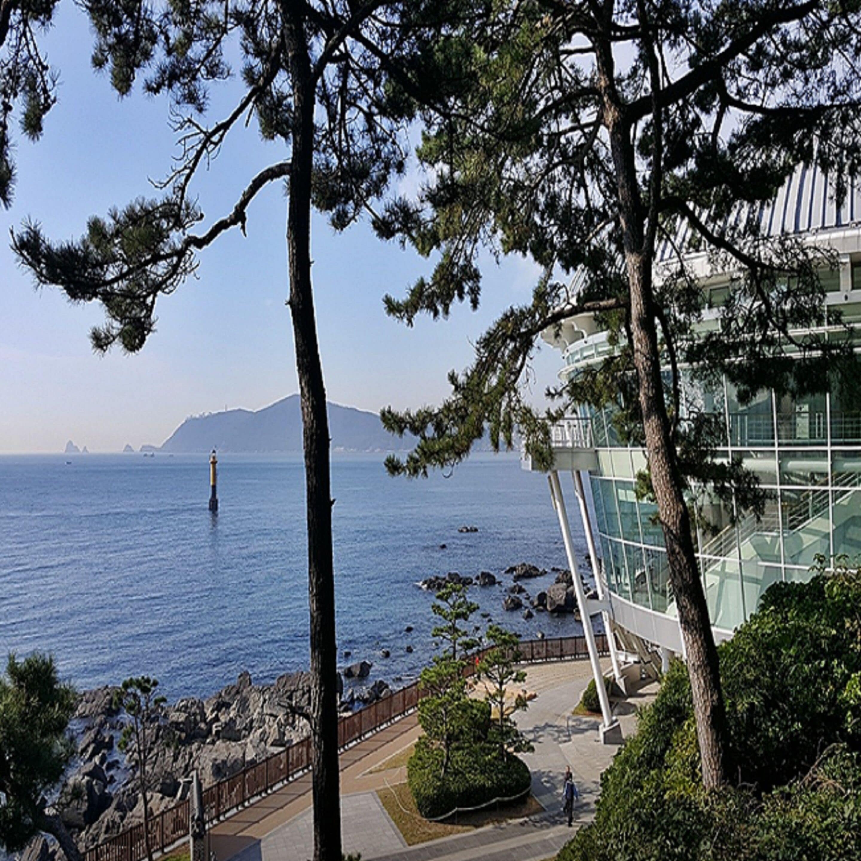 A coastal view with a modern building, a lighthouse in the distance, and a rocky shoreline.