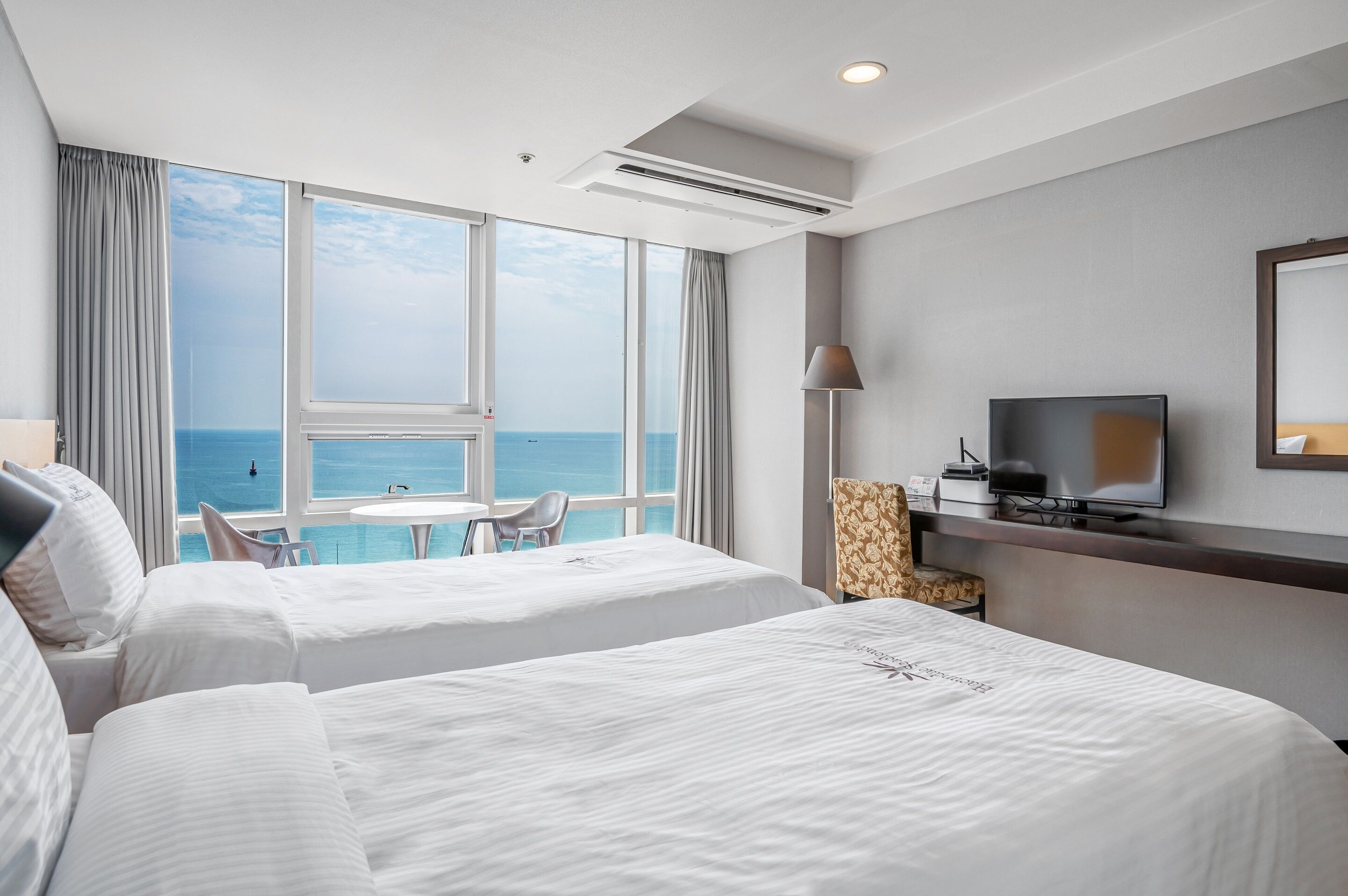 Habitación de hotel con dos camas, televisión, lámpara y vista al océano.