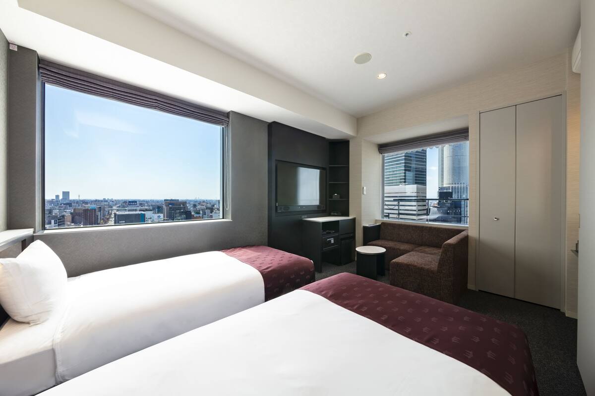Habitación de hotel con dos camas, un escritorio, una silla, un televisor y vistas a la ciudad.