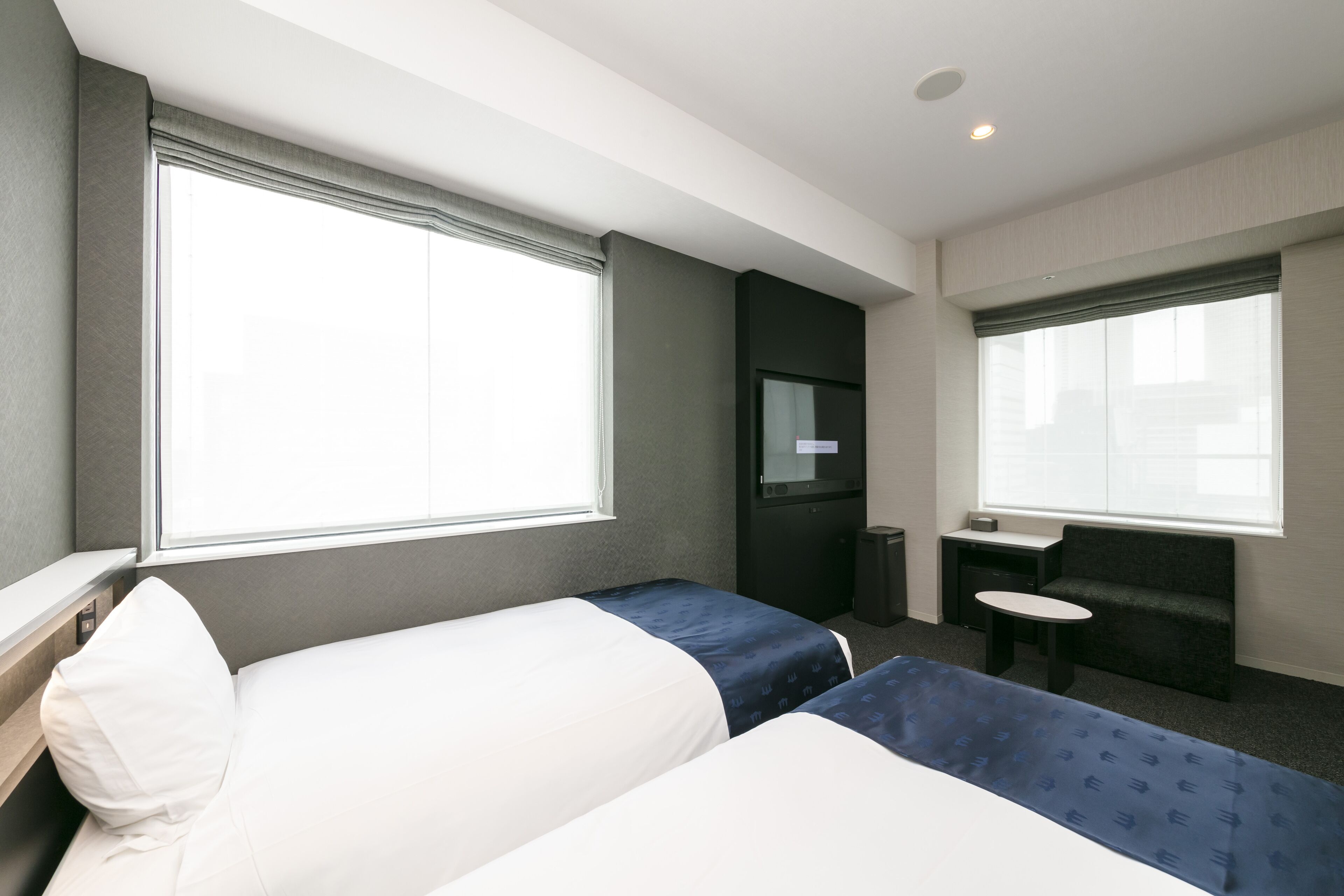A hotel room with two beds, a desk, a chair, and a window providing natural light.