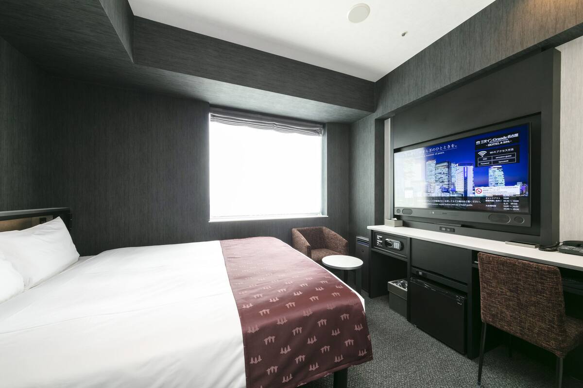 Una habitación de hotel moderna con una cama grande, un escritorio con televisor de pantalla plana, una silla y una kitchenette pequeña.