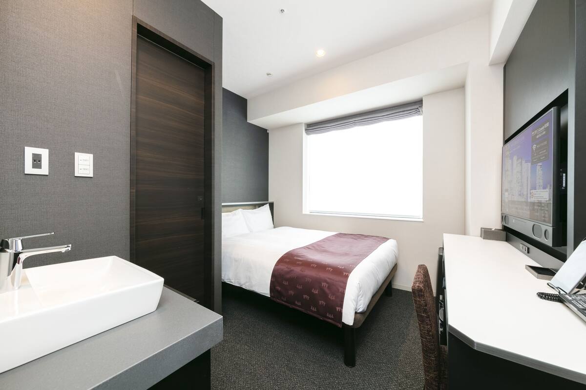 Una habitación de hotel moderna con una cama grande, una mesita de noche, televisión integrada y un baño con lavamanos y espejo.