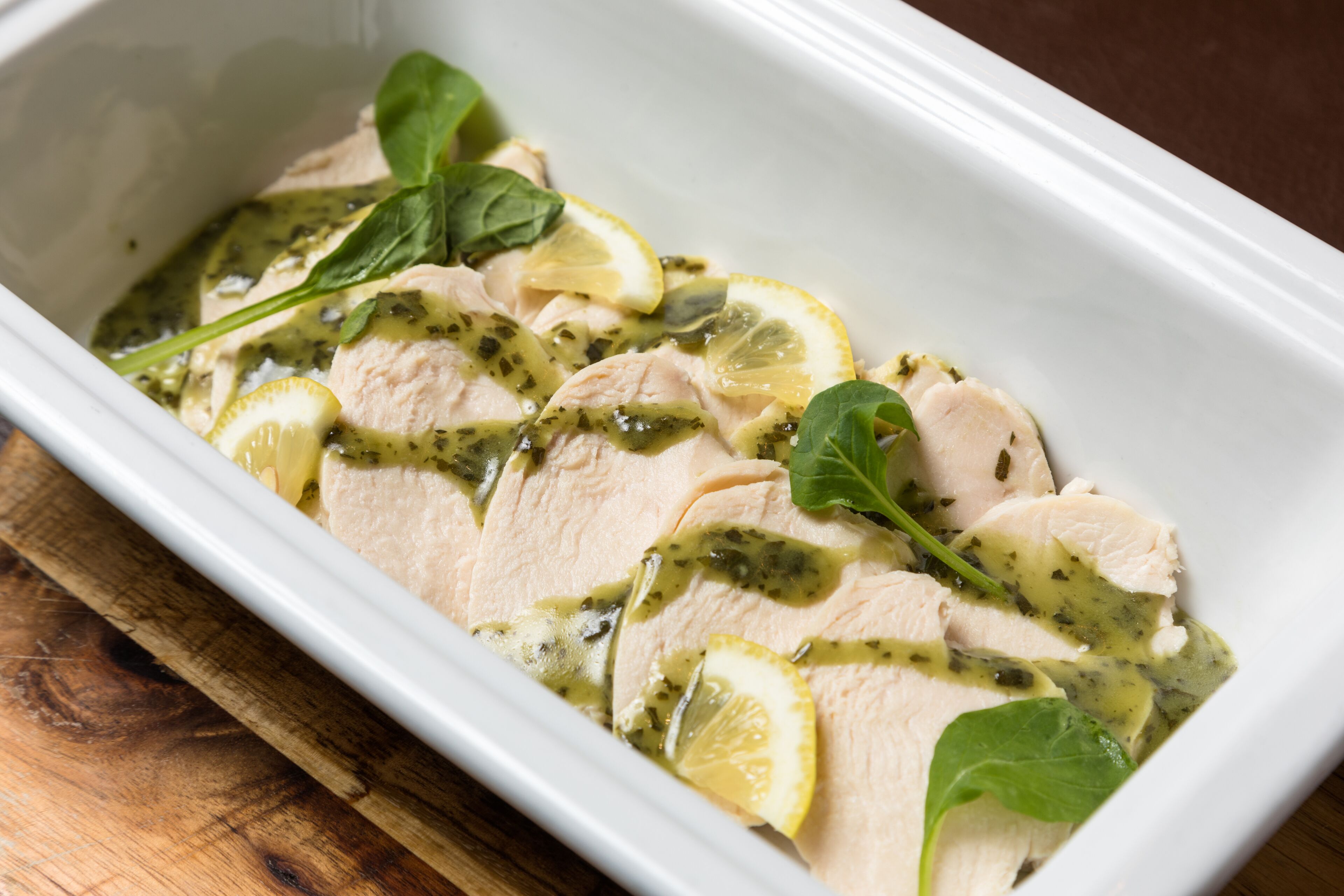 A white rectangular dish containing sliced chicken breast, lemon slices, and green leaves, likely spinach, in a sauce.