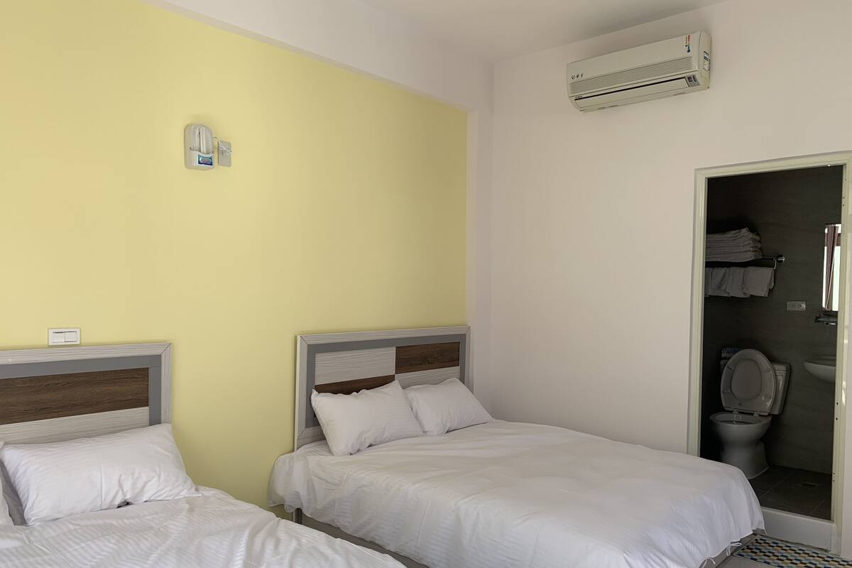 Een hotelkamer met twee bedden, een badkamer met toilet en wastafel, en een airconditioningunit aan de muur.