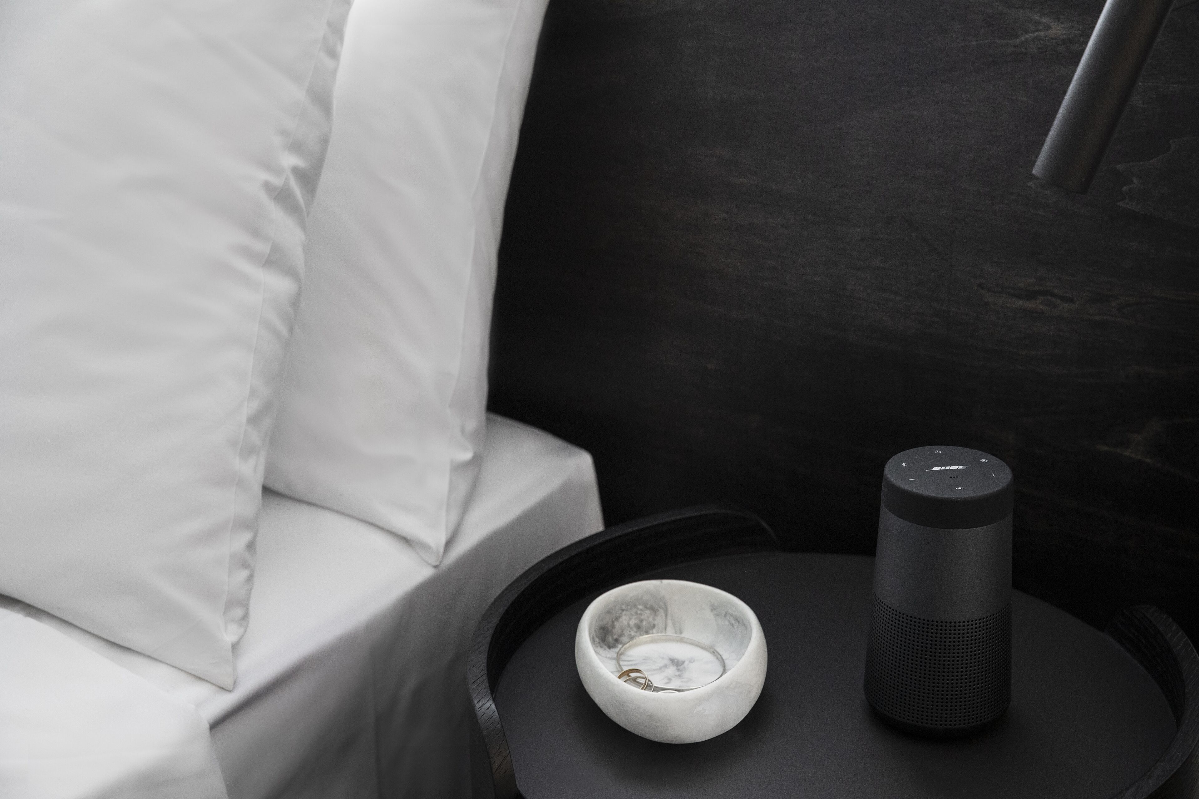 A modern room with a white bed, a small round table with a white bowl and a black speaker.