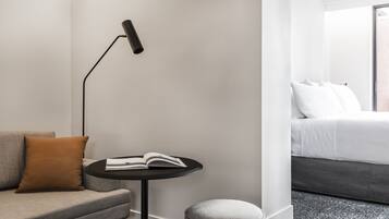 A modern hotel room with a sofa, a small round table with a book, a floor lamp, and a bed with white bedding.