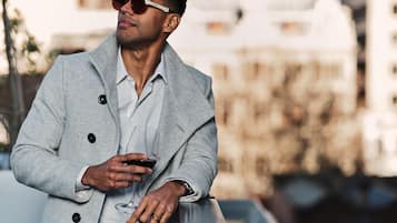 Una persona con un cappotto grigio tiene in mano un bicchiere, in piedi all'aperto con edifici sullo sfondo.