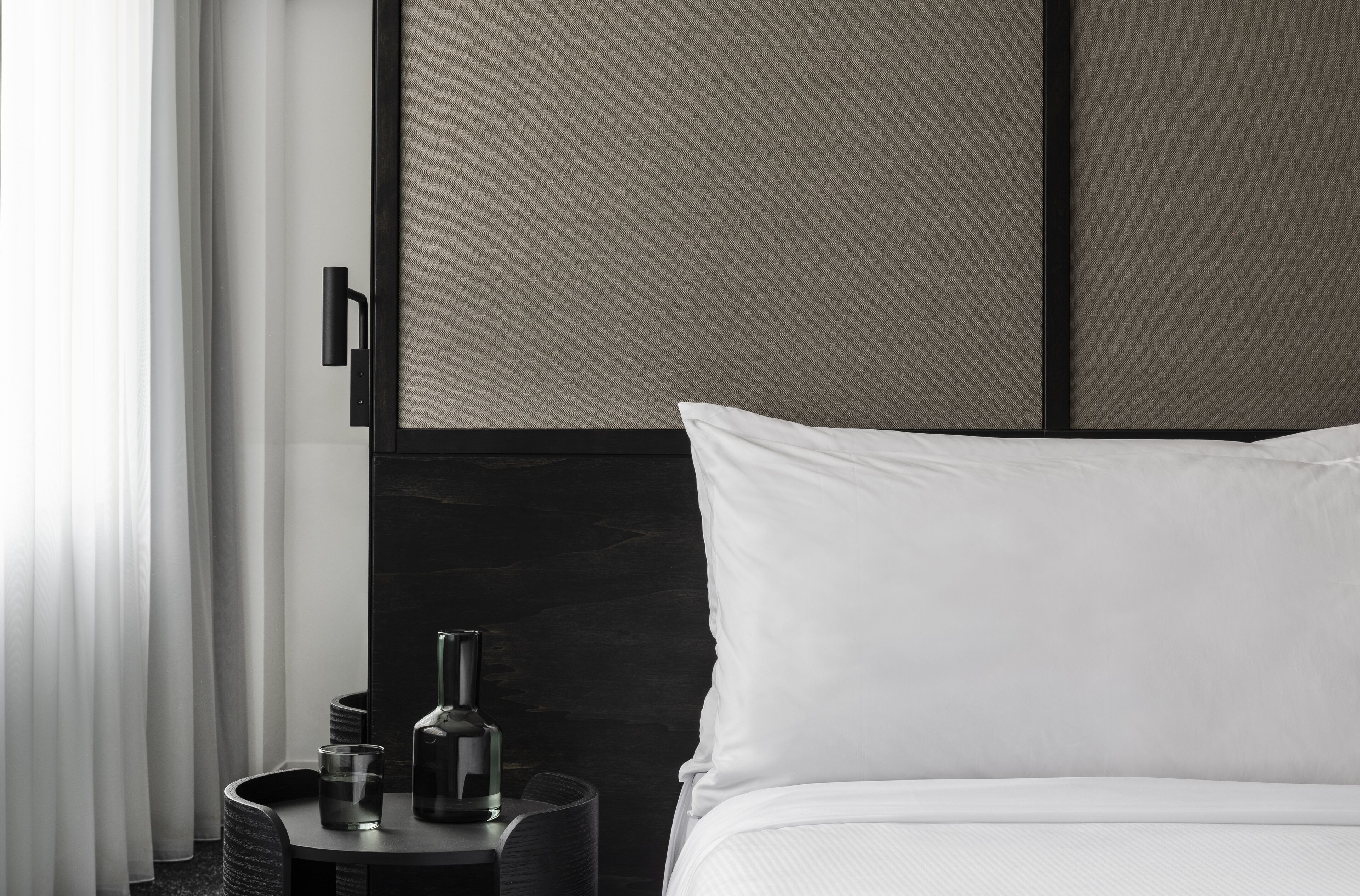 Una habitación de hotel moderna con una cama, una mesita de noche con una botella y vasos, y un panel en la pared.