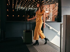 Een persoon in een mosterdkleurige outfit loopt met een koffer in een moderne, overdekte ruimte met een bord waarop staat 'Here's looking'.