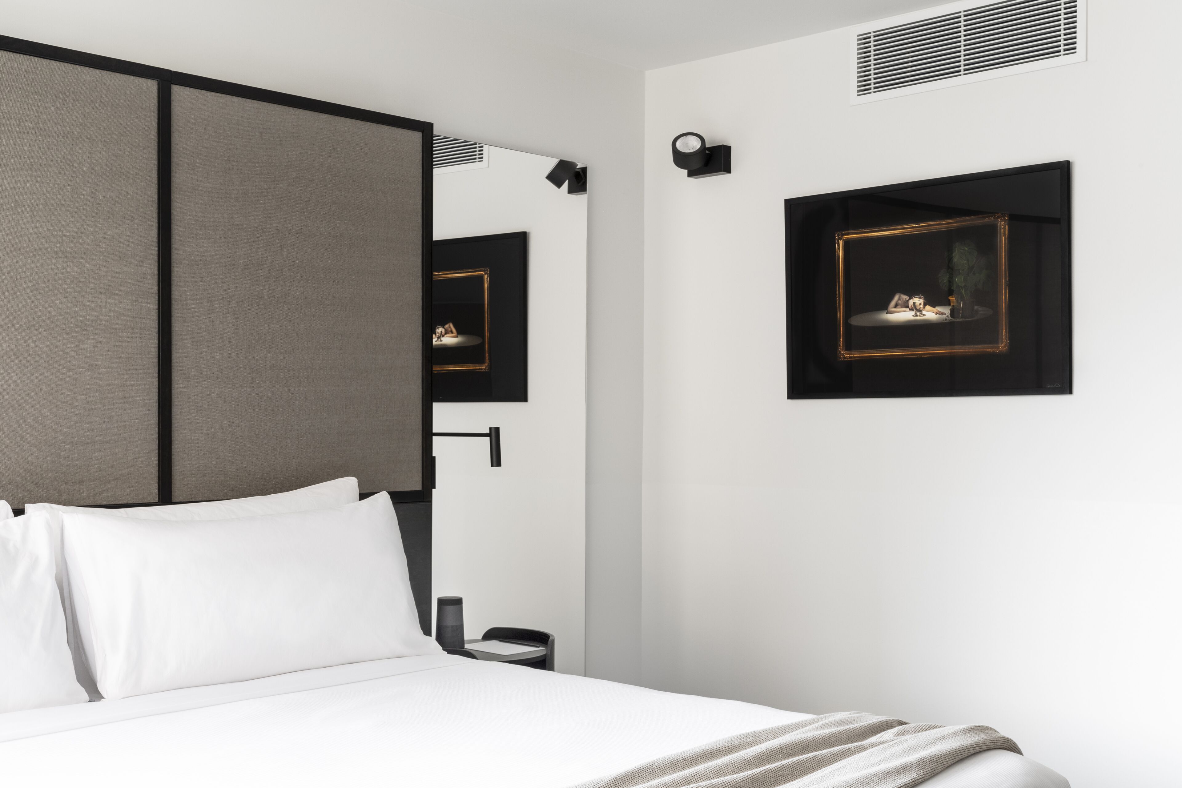 A modern hotel room with a bed, a bedside table, a lamp, and a framed picture on the wall.