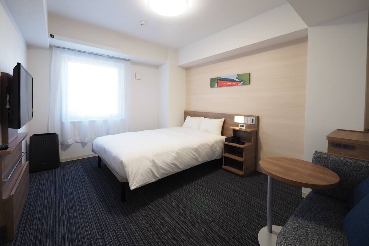A hotel room with a bed, a small desk, a chair, a TV, and a window with curtains.