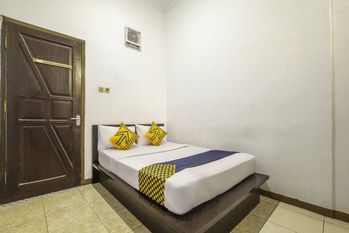 Hotelkamer met een grote houten deur, een bed met witte lakens en gele kussens met een patroon, en een kleine airconditioning op de muur.
