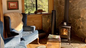 Ein gemütliches Wohnzimmer mit Steinwand, Holzofen, blauem Sofa und Fenster mit Blick auf Bäume.
