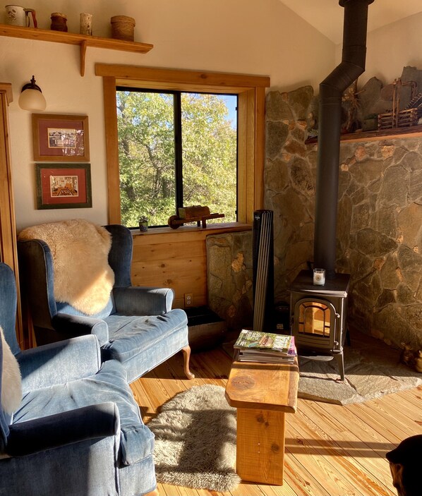 A cozy living room with a stone wall, a wooden stove, a blue sofa, and a window with a view of trees. - Private 40-acre Top of the Mountain Cabin with Stunning Panoramic Views, Deck (Julian)