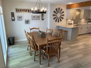 A dining area with a wooden table and chairs, a chandelier, and a kitchen with light-colored cabinets.