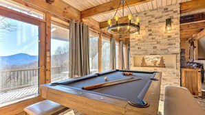 A pool table in a room with wooden beams, a chandelier, and a view of mountains through large windows. - Golf, Sauna, Creek: Spectacular Smoky Mtn-View Gem (Maggie Valley)