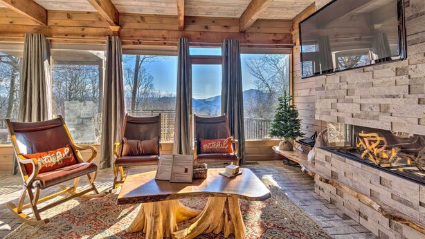 A cozy room with a fireplace, wooden furniture, and large windows offering a view of the outdoors. - Golf, Sauna, Creek: Spectacular Smoky Mtn-View Gem (Maggie Valley)
