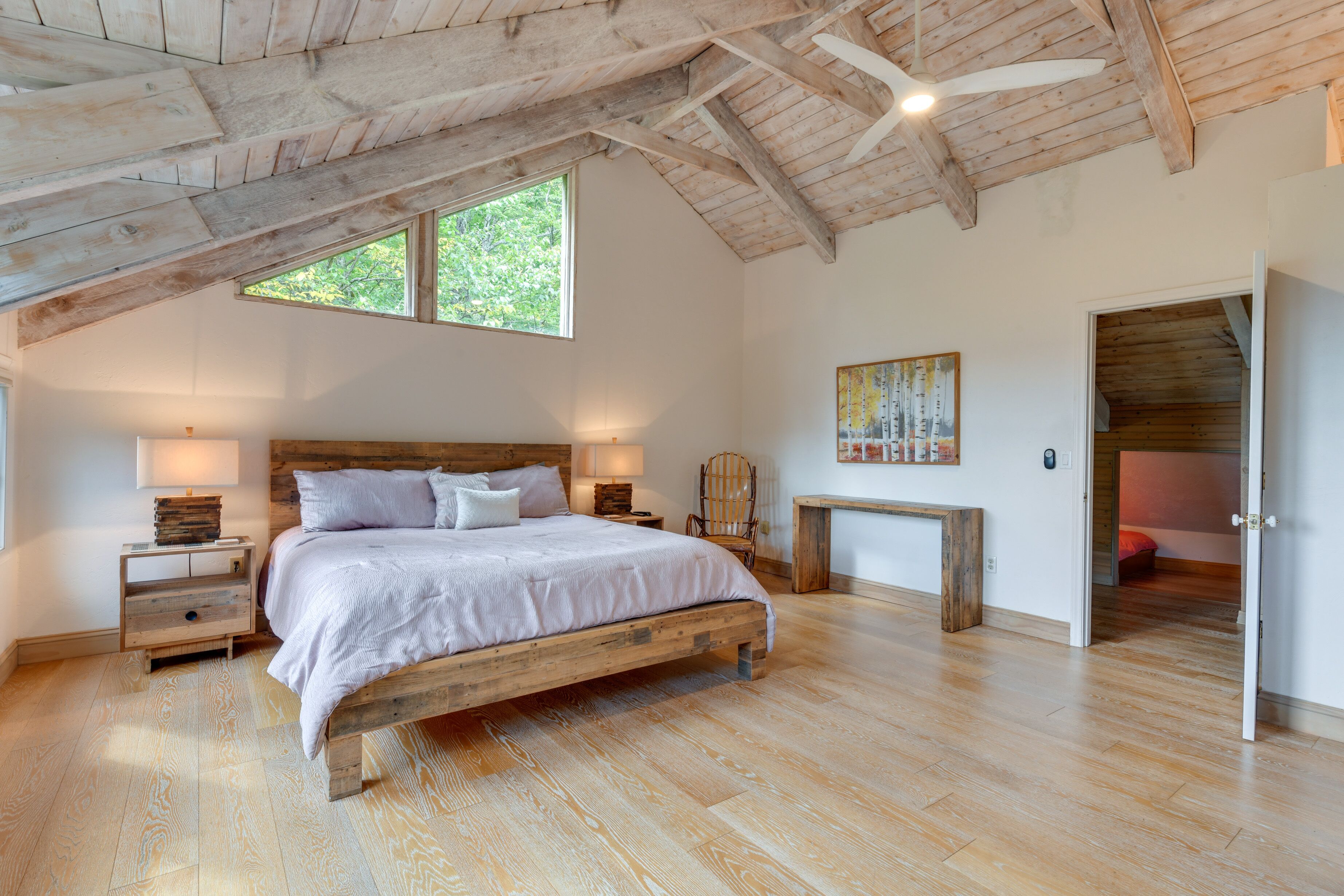 Un dormitorio amplio con una cama de madera, una mesita de noche, una silla y un escritorio. Hay una ventana que proporciona luz natural y un ventilador de techo.
