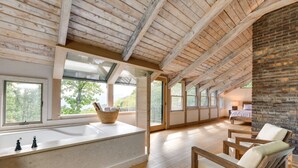 A spacious room with a wooden ceiling, large windows, a built-in bathtub, and a brick wall. - Golf, Sauna, Creek: Spectacular Smoky Mtn-View Gem (Maggie Valley)