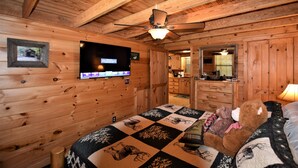 Ein Zimmer in einer Holzhütte mit einem Bett, einem Deckenventilator, einem Fernseher, einem Spiegel und einem Teddybär.