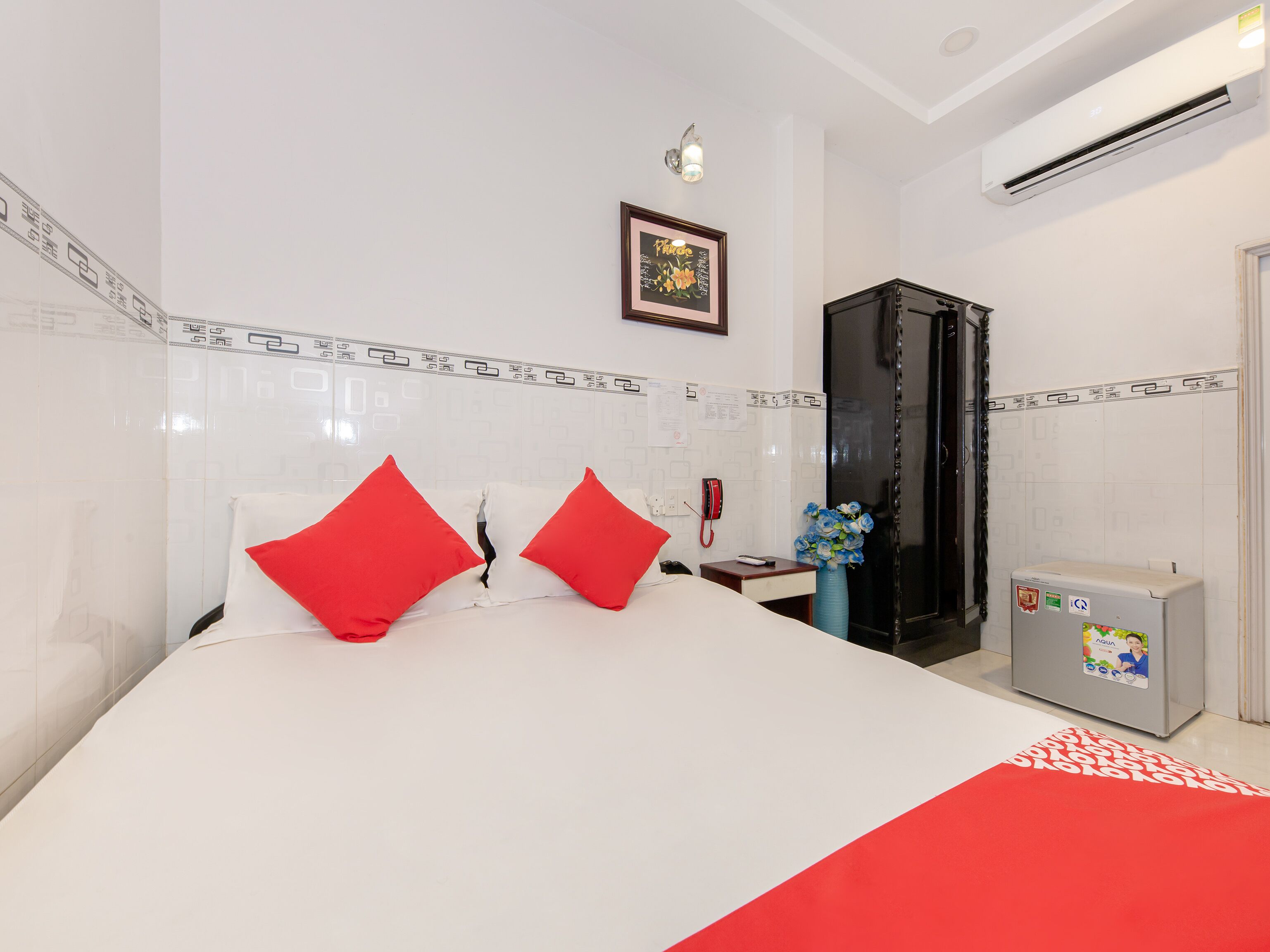 Una habitación de hotel con cama, mesita de noche, teléfono, un jarrón con flores y un equipo de aire acondicionado.