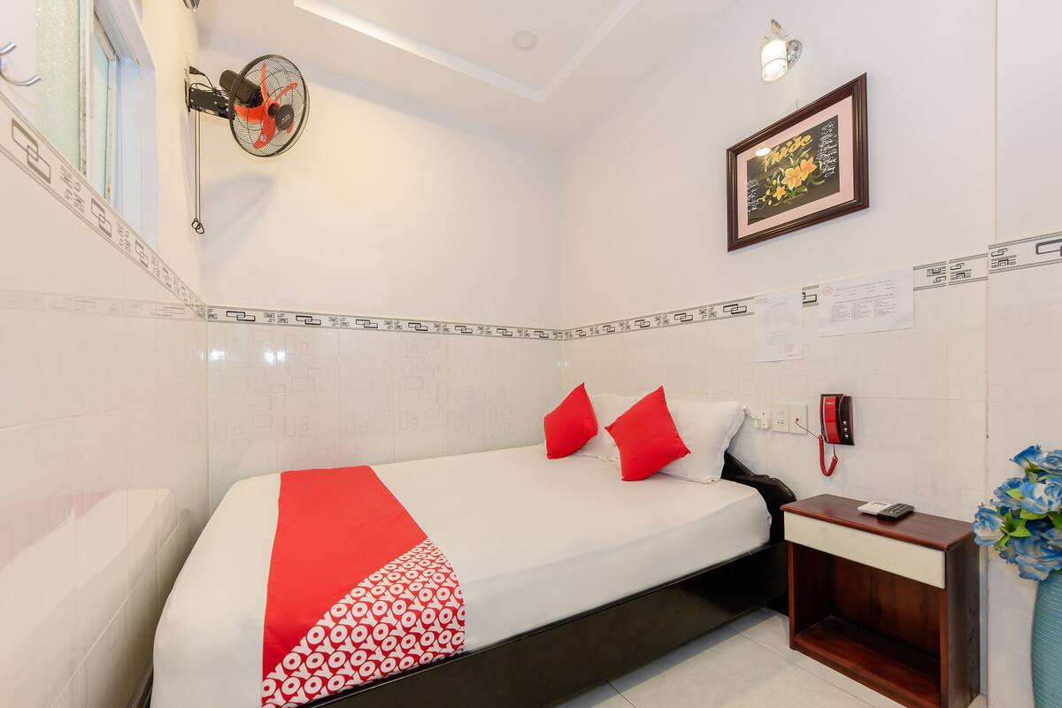 Une chambre d’hôtel avec un lit, une table de chevet, une commode, un ventilateur fixé au mur, un tableau encadré et un vase avec des fleurs.