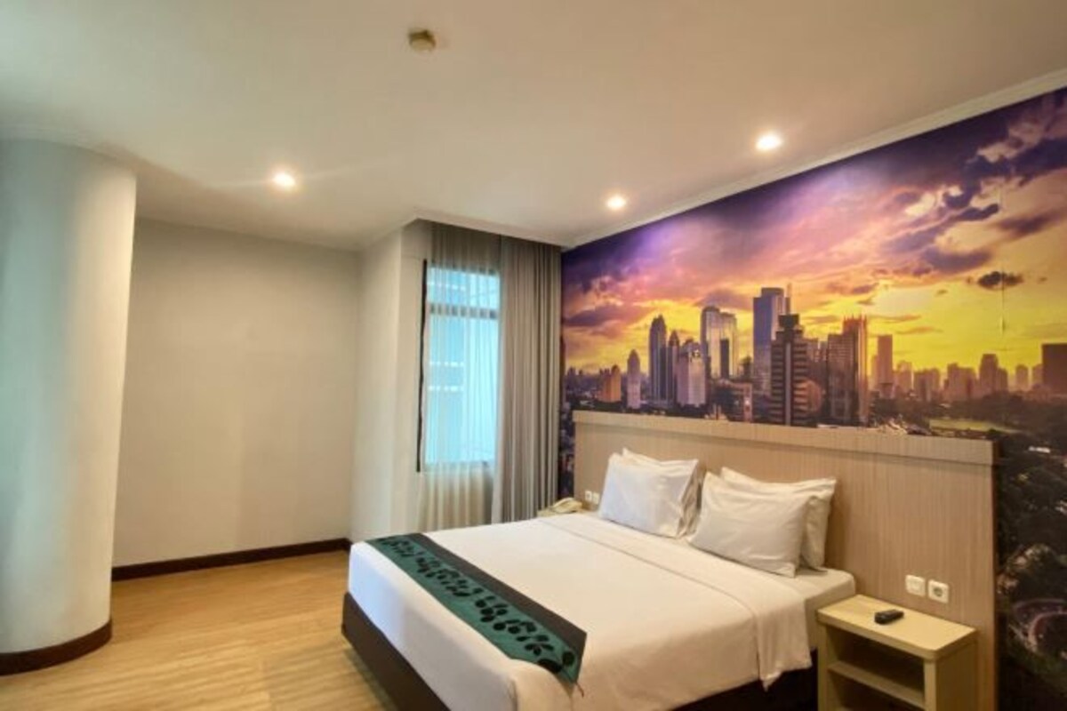 A hotel room with a bed, a nightstand, a window with curtains, and a cityscape mural on the wall.