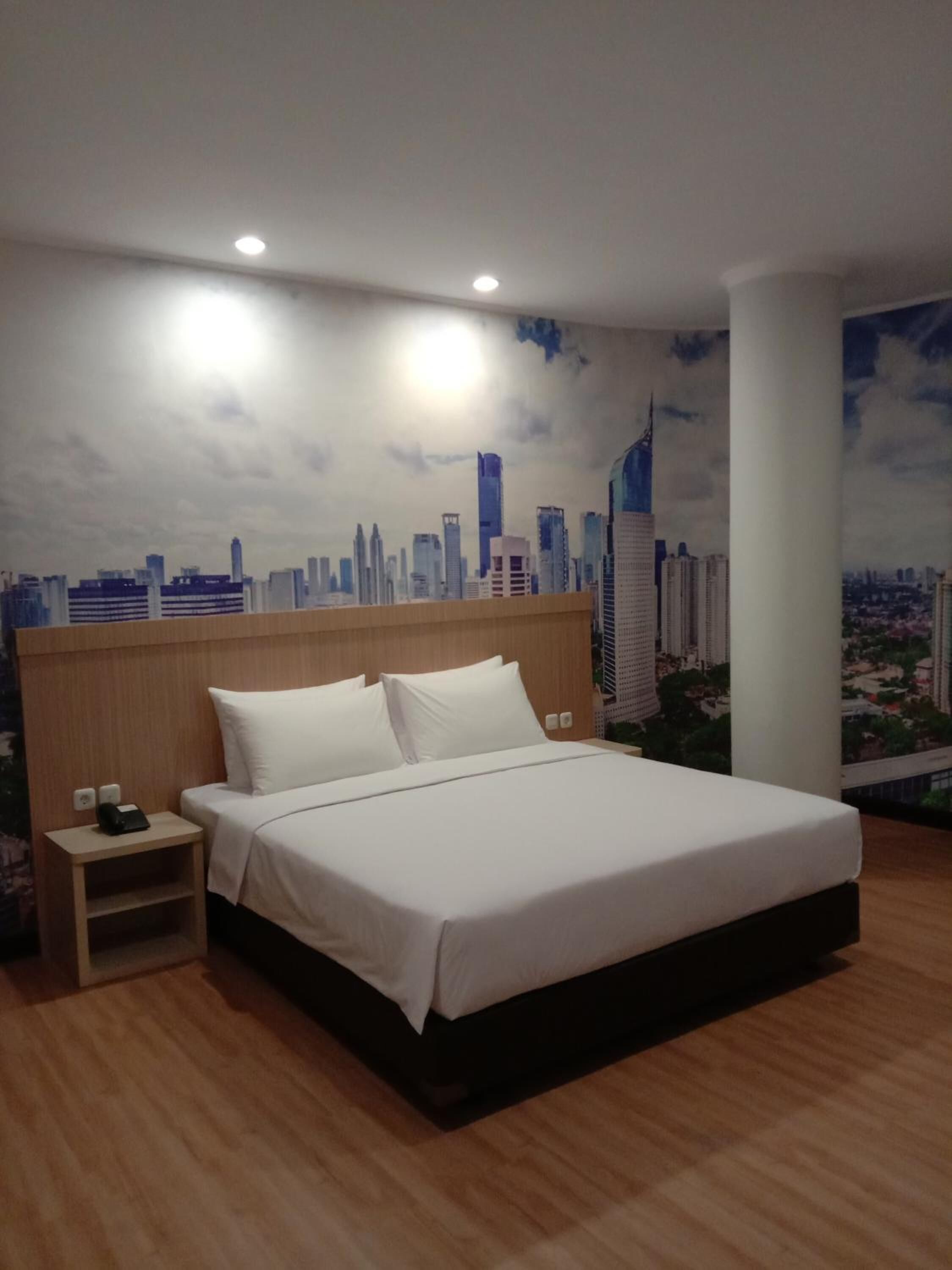 Une chambre d’hôtel avec un lit, une tête de lit en bois, une table de chevet et une fresque représentant un paysage urbain sur le mur.