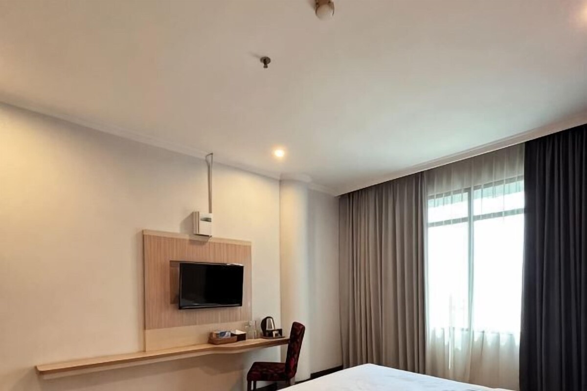 Habitación de hotel con cama, televisión montada en la pared, escritorio con silla y ventana con cortinas.
