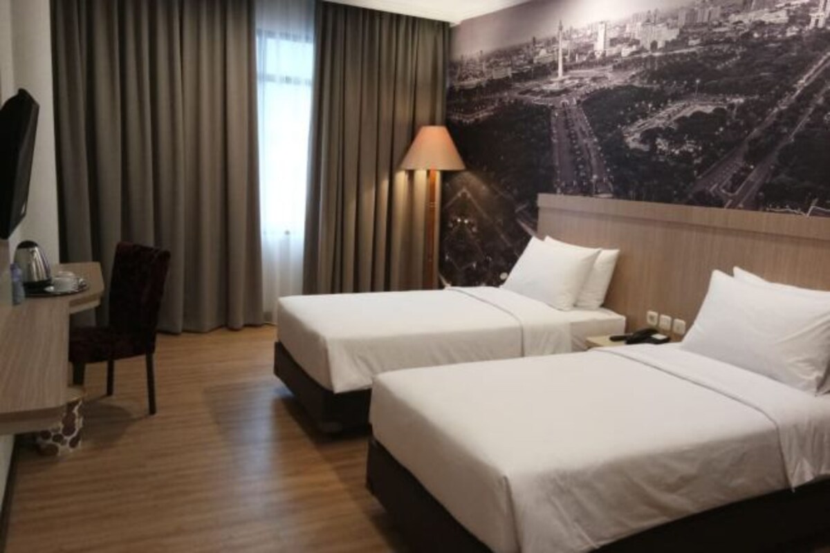 A hotel room with two beds, a desk, a chair, a lamp, and a cityscape mural on the wall.