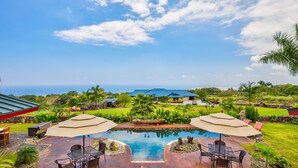 A pool area with two umbrellas, chairs, and a view of the ocean. - The “Promise Tree” is a great family  gathering place in Kailua Kona! (Kailua-Kona)